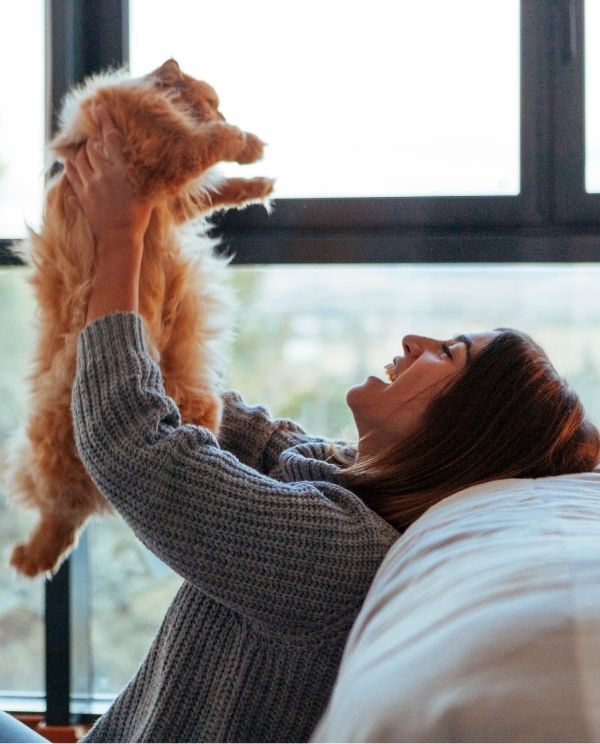 A person in a grey sweater holds up a fluffy orange cat while lying on the floor next to a bed.