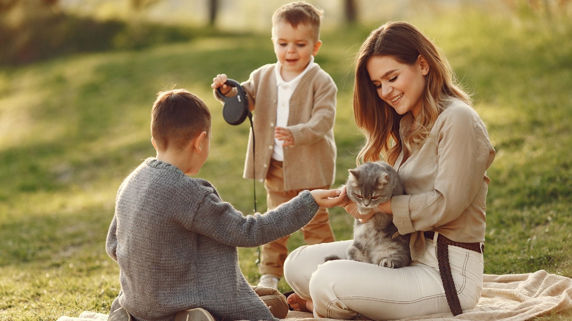 A woman sits on a blanket in a grassy field, holding a cat. Two children are nearby, one petting the cat and the other holding a retractable leash.